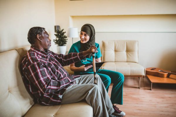 A Muslim female doctor is visiting her older patient of Afro-American ethnicity. An Arabian female healthcare worker is helping her older male patient in the living room at home.